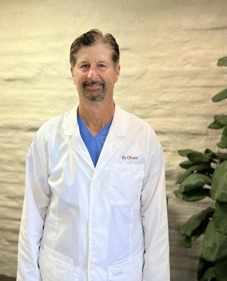 Dentist John Olson DDS in scrubs and white coat in dental office in Huntington Beach, CA